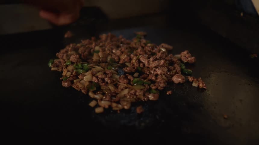 A chef stirs and cooks chopped meat with onions and peppers on a large, hot flat-top griddle
