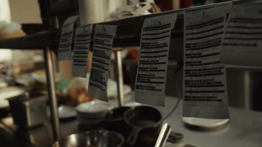 A slow-motion dolly-in shot reveals a line of order tickets hanging in a busy restaurant kitchen