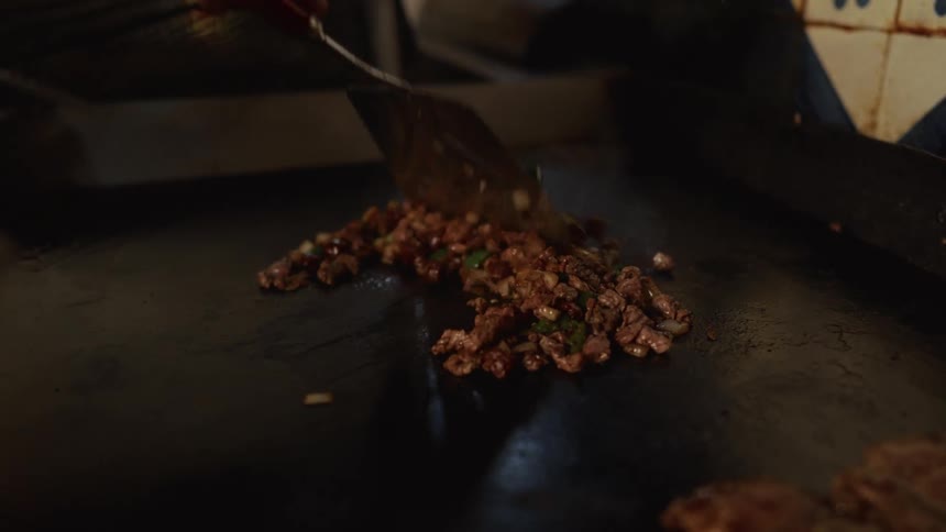 A chef stir-fries chopped meat and vegetables with a spatula on a large hot griddle in slow motion