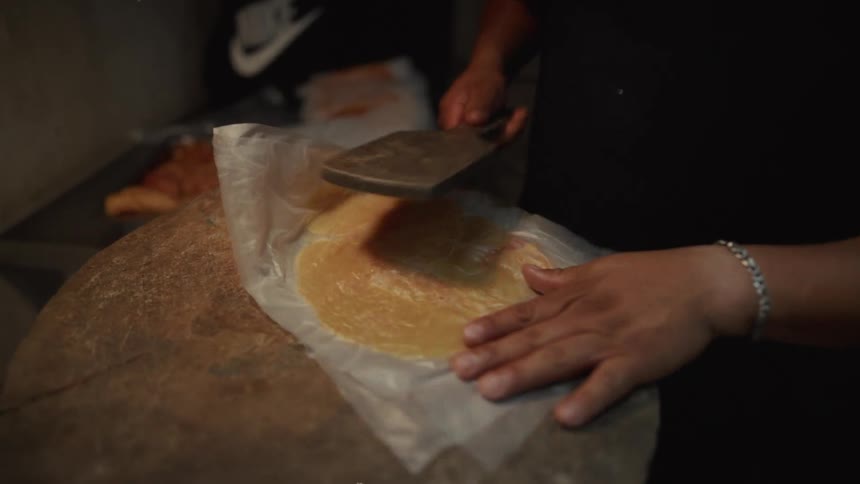 A cook pounds and flattens a raw chicken breast with a meat tenderizer on a large wooden block