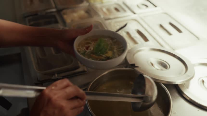 Slow motion shot of a chef in a restaurant kitchen carefully ladling hot broth into a bowl of chicken noodle soup topped with avocado