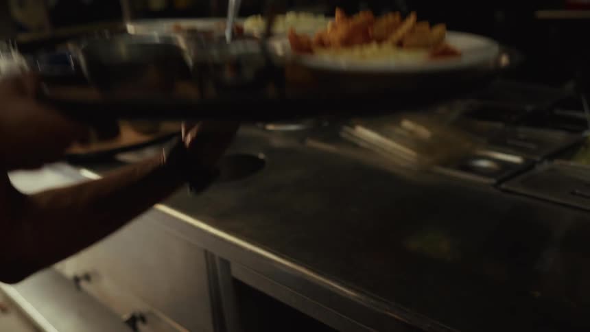 A waiter in a dimly lit restaurant kitchen carefully picks up a large tray filled with freshly prepared meals in slow motion