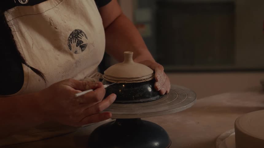 An artisan potter carefully smooths and carves decorative lines into a handmade clay lid using a tool in her workshop
