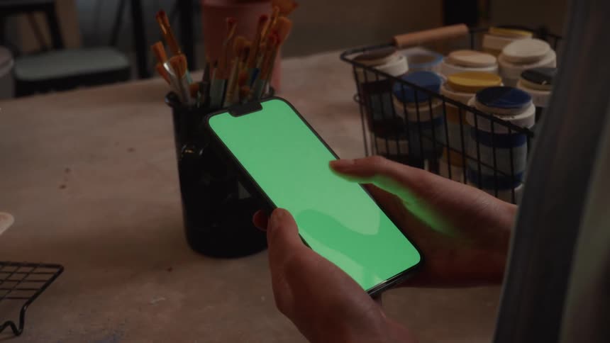 An artist's hands scroll and tap on a smartphone with a green screen while sitting at a table with paint and brushes