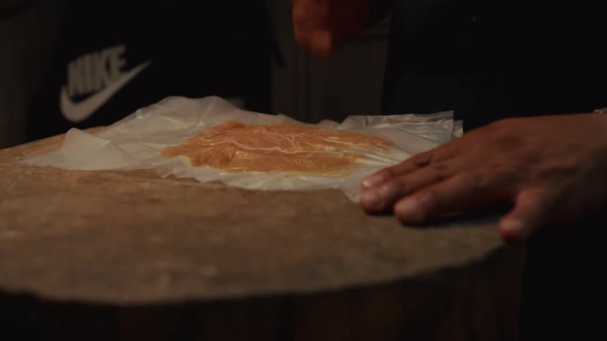 A chef in a dark kitchen uses a metal meat tenderizer to pound a piece of chicken flat in slow motion