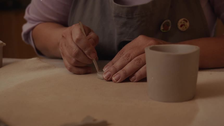 An artist meticulously uses a sculpting tool to score a circular piece of clay in a cozy, warm-lit studio