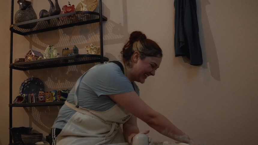 A cheerful woman wearing an apron joyfully shapes a piece of pottery on a wheel in a cozy studio