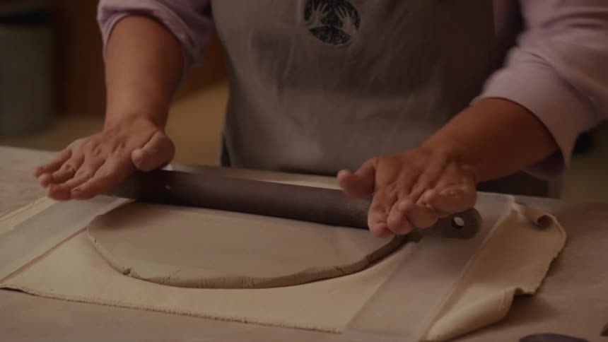 A ceramic artist wearing an apron uses a rolling pin to carefully flatten a slab of clay in their workshop