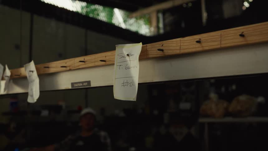 A person's hand places a handwritten food order ticket onto a service rack in a commercial kitchen in slow motion