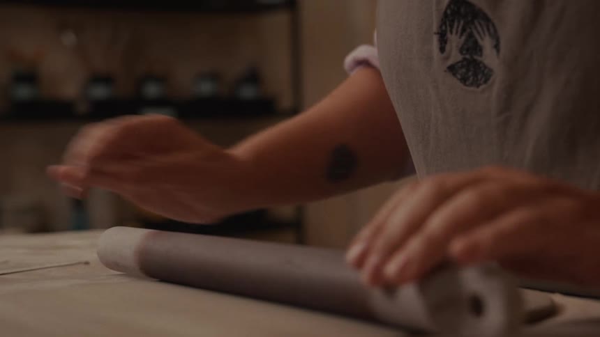 A ceramicist rolls out a slab of clay with a rolling pin and then begins to trim it with a needle tool in their studio