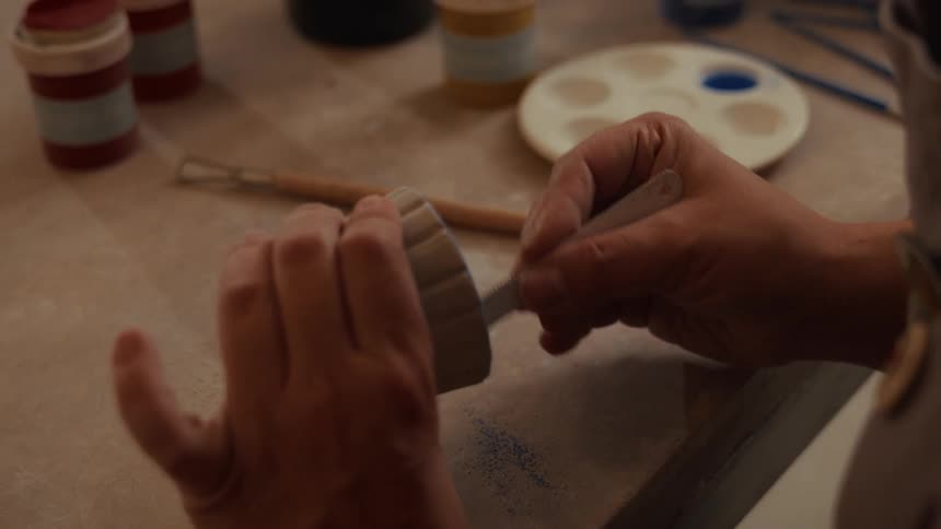 Over the shoulder shot of an artisan's hands meticulously carving details into a handmade clay cup at a workbench