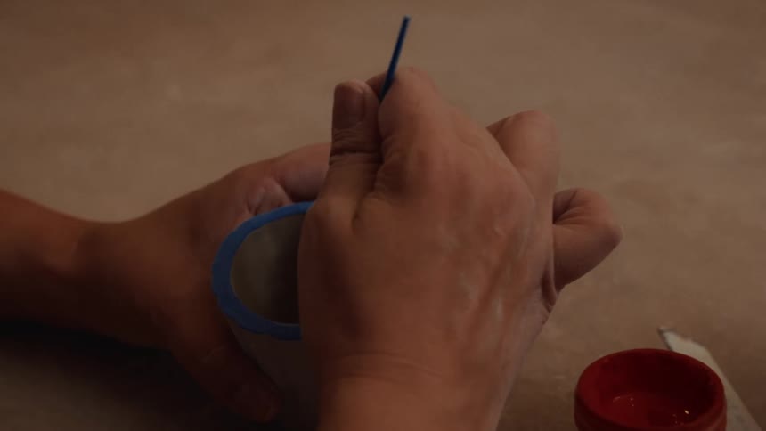A close-up view of an artisan carefully painting the inside of a scalloped ceramic cup with a fine brush and red paint