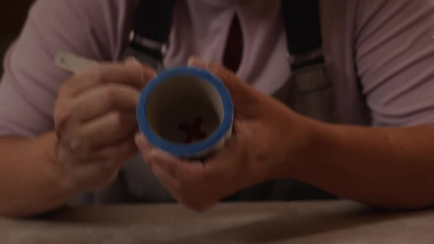 An artisan in an apron meticulously scrapes designs onto the side of a small handmade pottery cup in her workshop