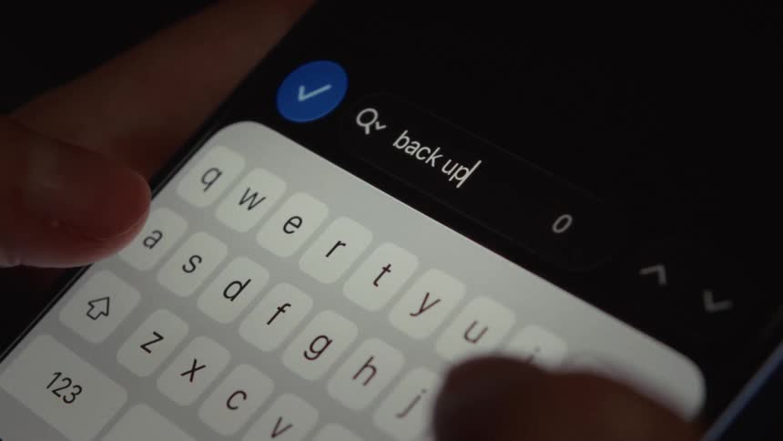 A close-up shot of a person typing the words 'back up' into a search bar on a smartphone in a dark room