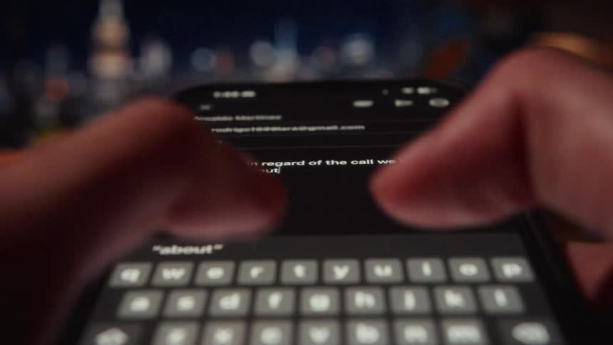 A person types a business email on a smartphone keyboard in a dark room with a blurry background