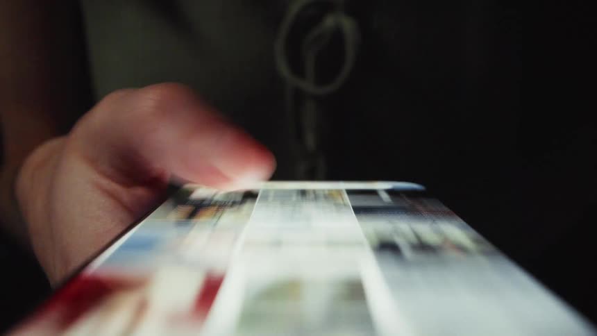 A macro close-up shot of a thumb rapidly scrolling through a social media photo feed on a smartphone in the dark