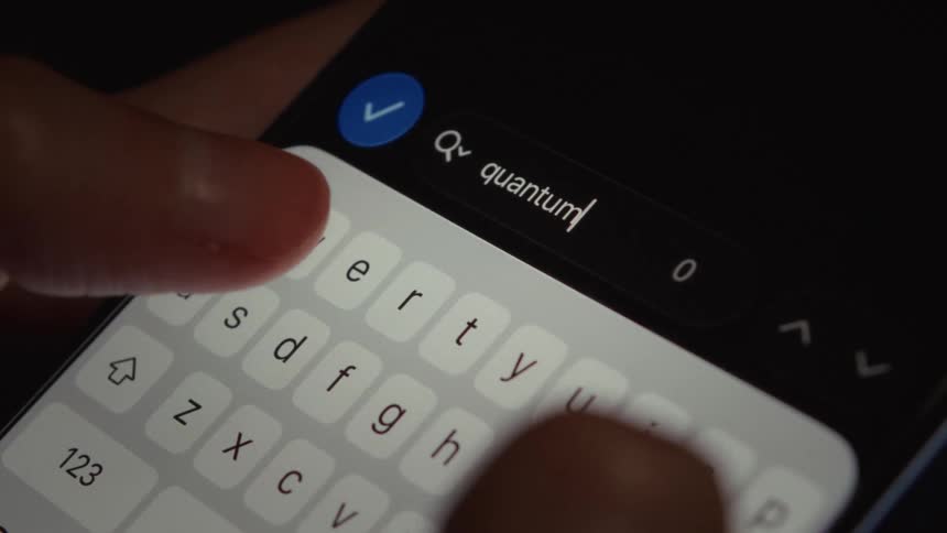 Fingers type the word 'quantum' into the search bar on a smartphone in a dimly lit room