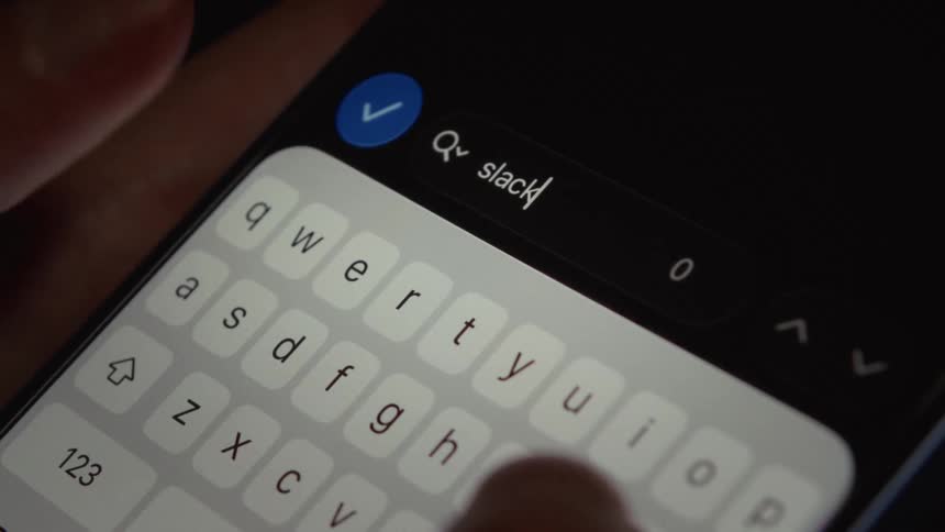 A person's fingers type the word 'Slack' into a search bar on a smartphone
