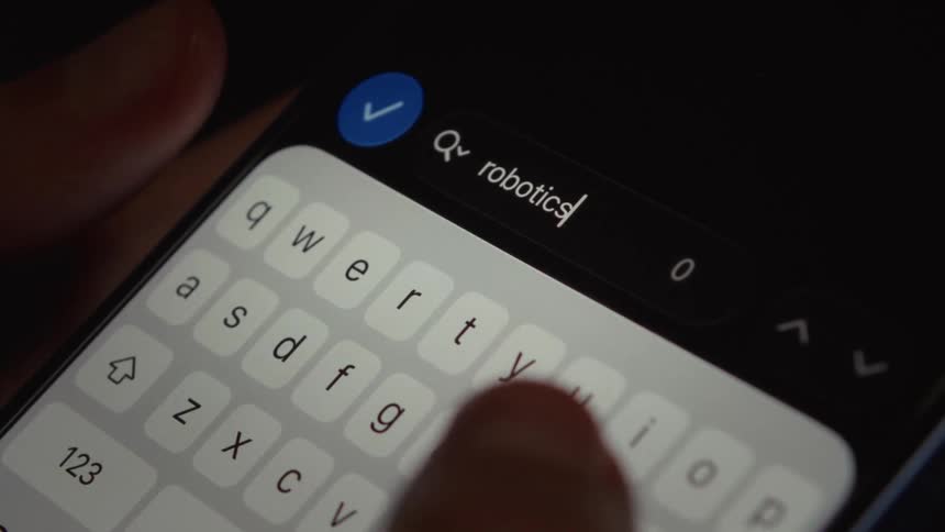 A person types the word 'robotics' into the search bar of a smartphone in a dark room