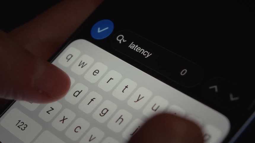 A close-up view of fingers typing the word 'latency' into a search bar on a smartphone screen in a dimly lit setting