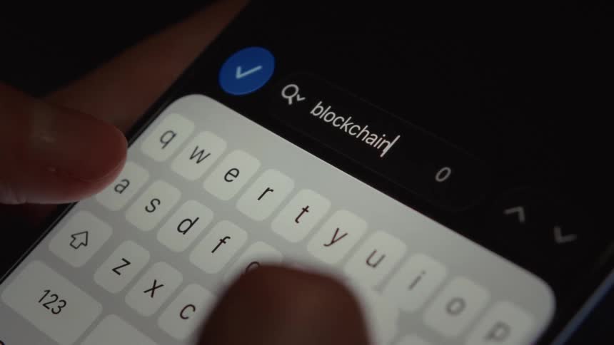 A close-up shot of fingers typing the word 'blockchain' into a smartphone's search bar in a dark room