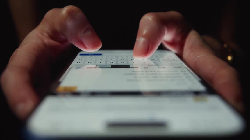 A dramatic low-angle close-up shot shows hands typing a message on a smartphone in the dark