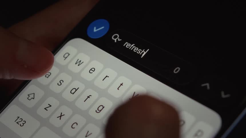 Close up of fingers typing the word 'refresh' into a search bar on a smartphone keyboard in a dark room