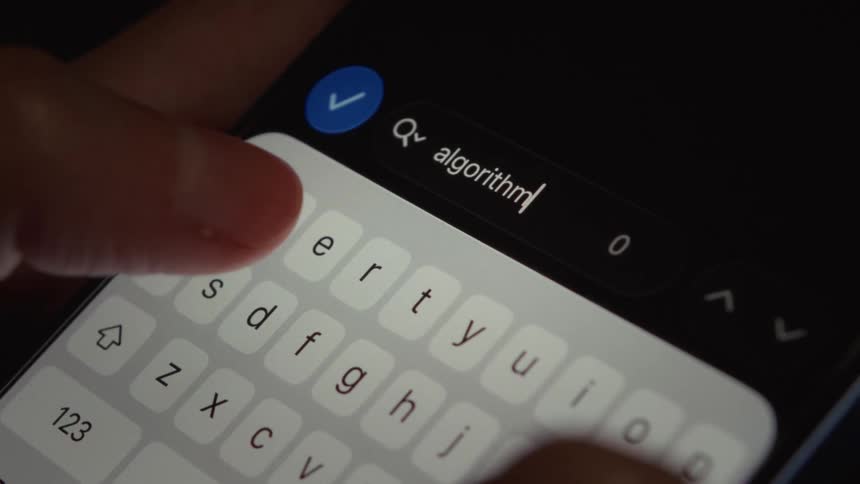 A close-up shot of fingers typing the word 'algorithm' into a search bar on a smartphone screen in dark mode