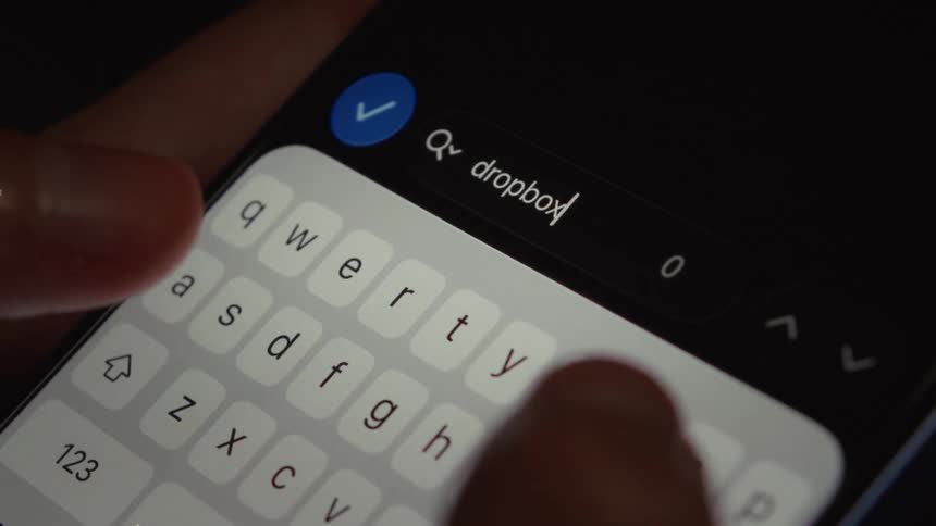 A close-up of a person typing 'dropbox' into a search bar on a smartphone in dark mode
