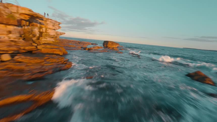 A fast-paced FPV drone flight speeds low over the ocean along a rocky shoreline bathed in the warm light of golden hour