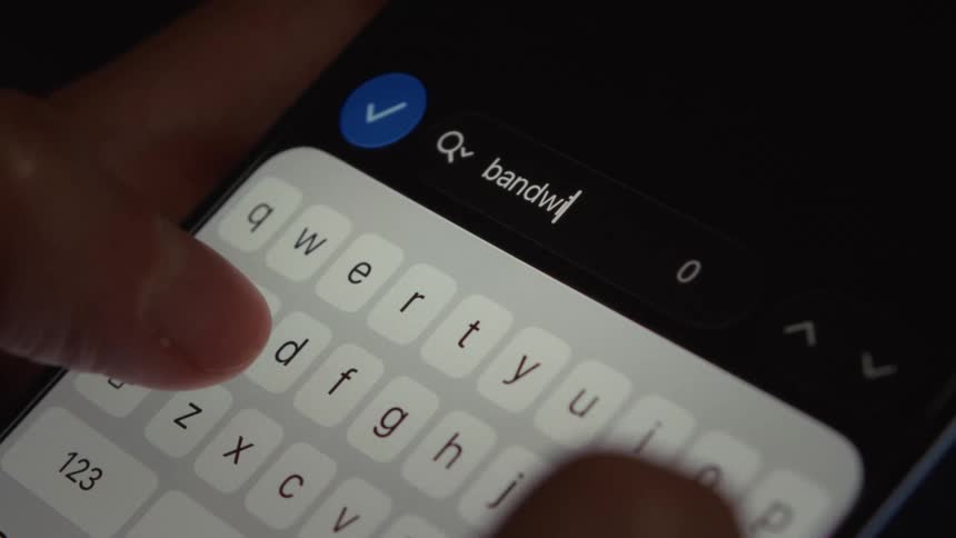 A close-up of fingers typing 'bandwidth' into a smartphone's search bar in a dimly lit room