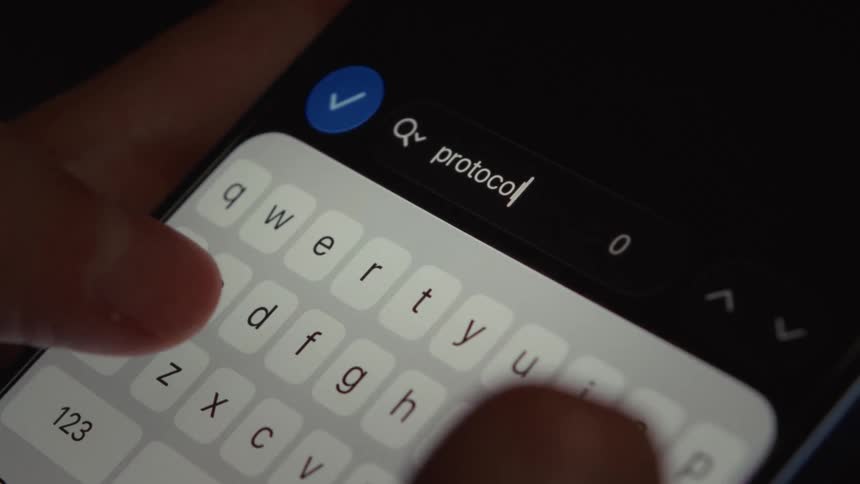 Extreme close-up of fingers typing the word 'protocol' into a search bar on a smartphone in a dark room