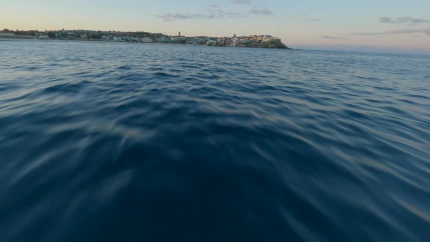 A fast-paced, low-angle drone shot skims across the surface of the deep blue ocean towards a distant coastal city skyline at sunrise