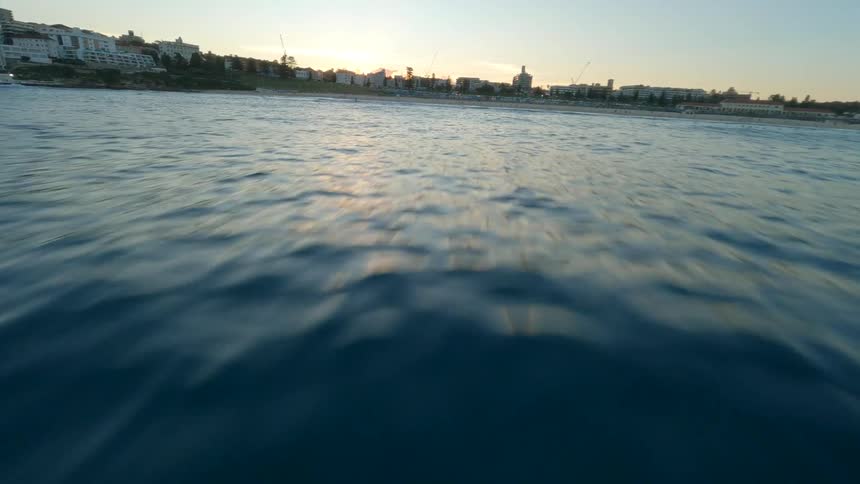 A dynamic FPV drone shot skims low over the ocean's surface towards a city coastline at sunset, capturing a person on an eFoil