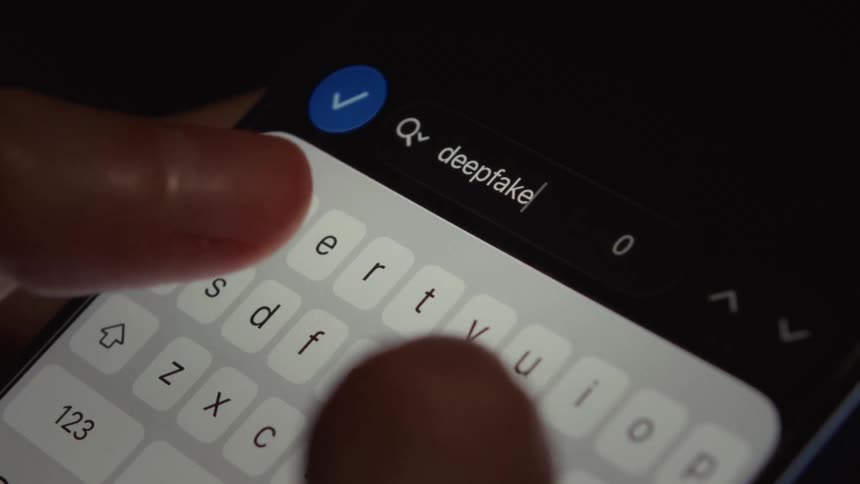 A close-up shot shows a finger slowly typing the word 'deepfake' into a search field on a smartphone in the dark
