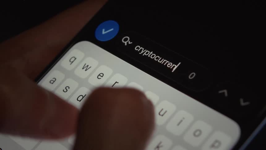 Close-up of fingers typing 'cryptocurrency' into a search bar on a smartphone screen in a dimly lit room