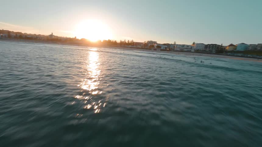 An FPV drone soars low over the rocky coastline and out to sea, capturing surfers riding waves under the beautiful golden sunset