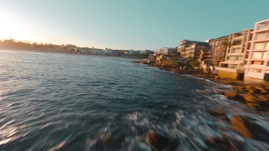 An exhilarating FPV drone speeds along a rocky coastline at golden hour, capturing the sun's reflection on the ocean waves crashing ashore