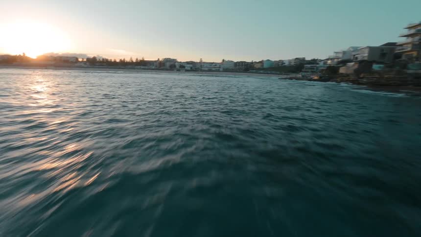 An immersive FPV drone speeds over the ocean along a rocky coastline and towards a beach town at sunset