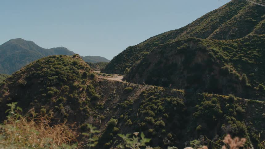 A rapid pull-back shot reveals a winding road curving through lush green mountains on a bright, sunny day