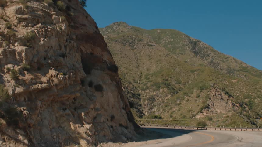 A fast zoom-out shot from a rocky cliff reveals a winding mountain road on a sunny day