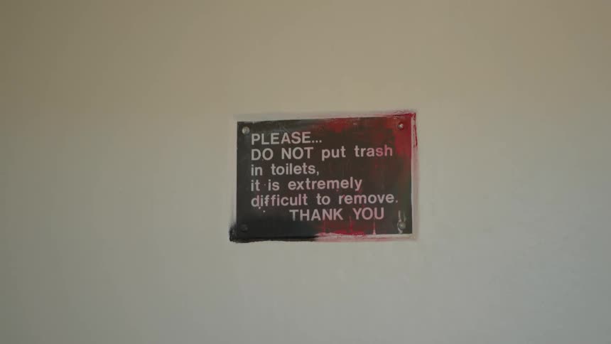 A stark public toilet is shown before the camera focuses on a worn sign warning against putting trash in it