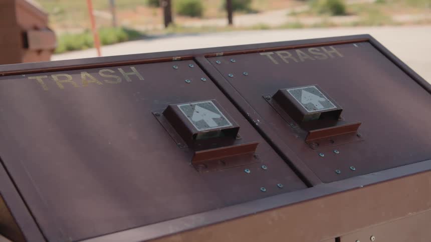 A close-up shot of a brown, double-lidded trash receptacle at an outdoor recreational area