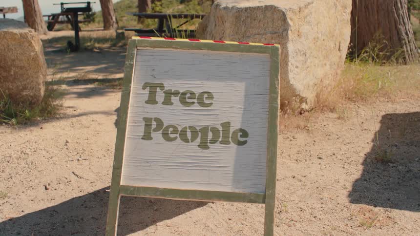 A weathered wooden sign for the 'Tree People' organization stands in a sunny park with tall trees and picnic tables