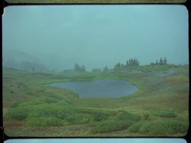 Vintage 16mm film footage of rain falling on a small, secluded mountain lake on a foggy, overcast day