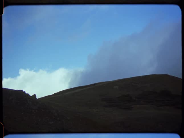 Distant figures hike along a shadowy mountain ridge under a vast, cloudy sky