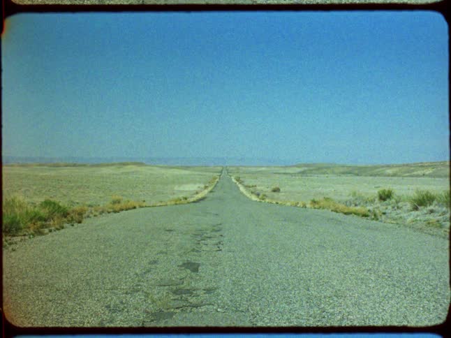 Super 8 film captures a fast zoom-in on a long, lonely road stretching across a vast, empty desert landscape under a clear blue sky