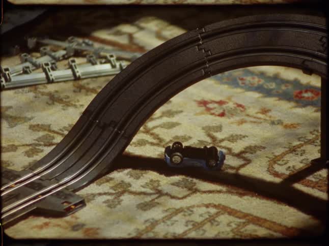 Retro-style home movie footage shows toy slot cars racing on a track while a blue car is crashed nearby on the floor
