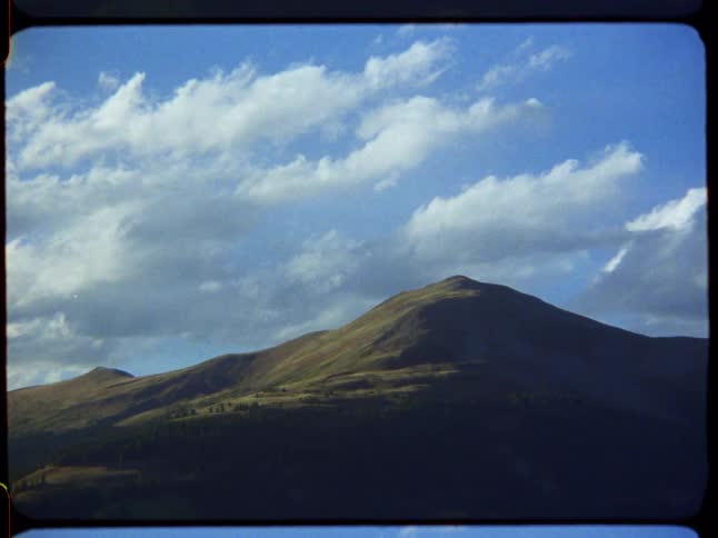 A vintage film shot pans up from a majestic, rolling mountain to a vast blue sky dotted with white clouds