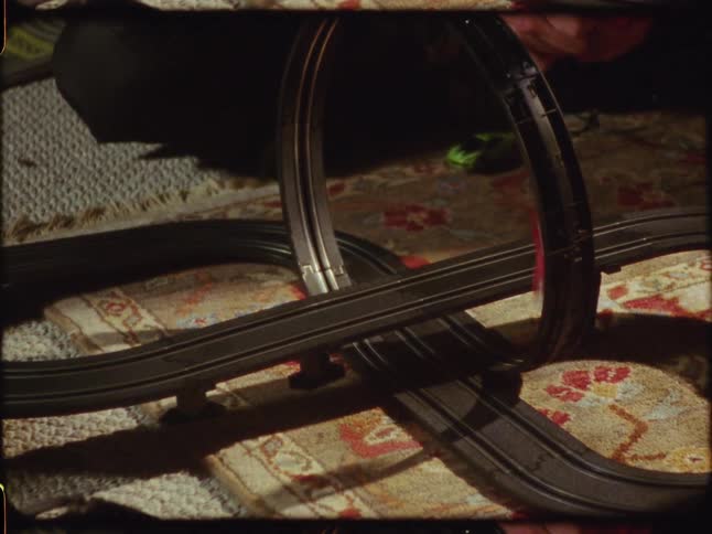 Vintage home movie footage shows two colorful slot cars racing around a loop track on a living room rug